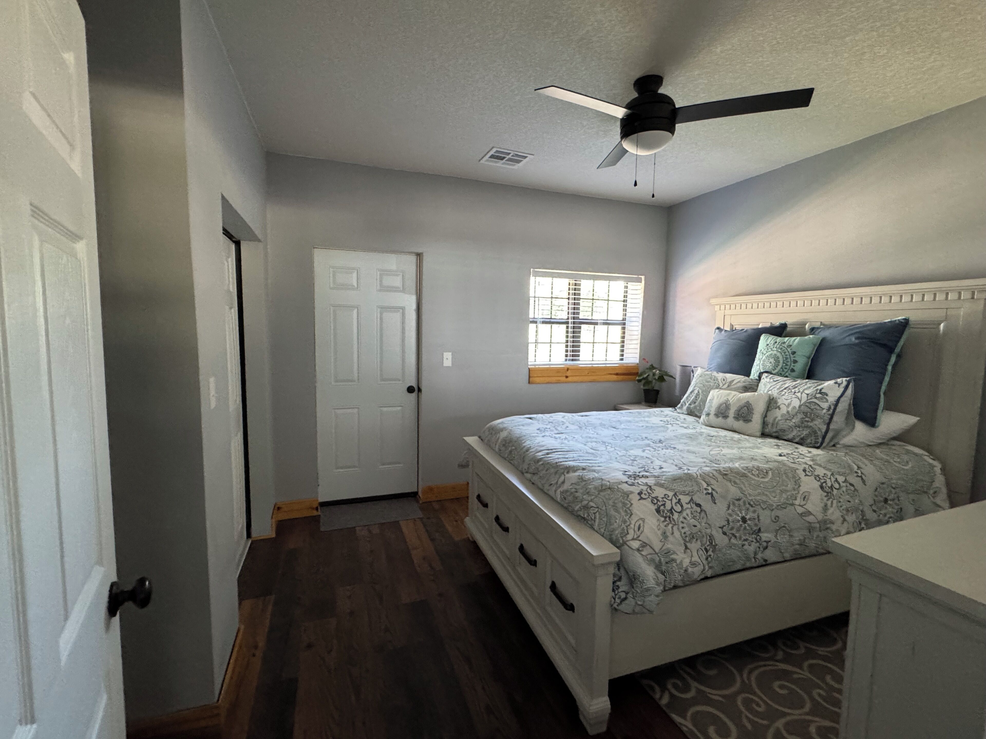 A bedroom with a bed, a nightstand, a window with blinds, a ceiling fan, and a door.