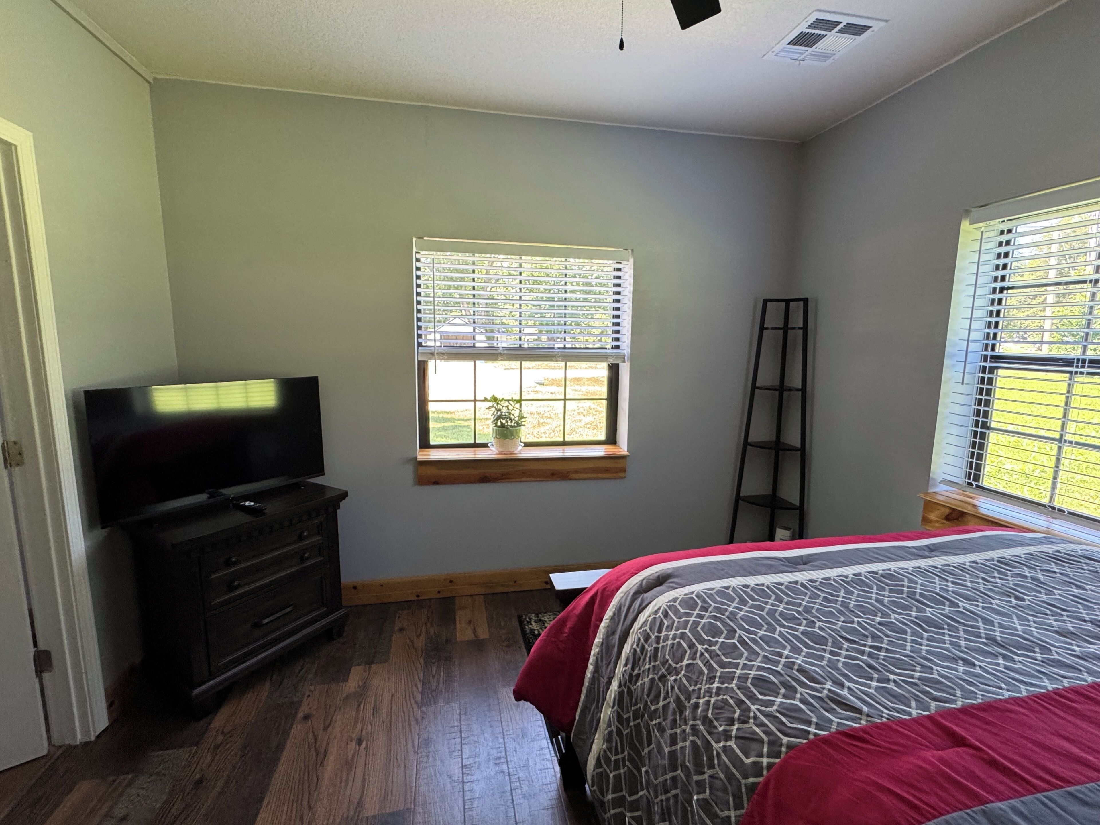 A bedroom with a TV, a window with blinds, a wooden floor, and a bed with patterned bedding.