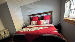 A bedroom with a bed, a nightstand, a window with blinds, and a wall-mounted shelf.