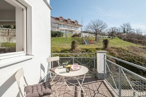 Un balcone con tavolo e sedie, una poltrona imbottita e vista su una zona residenziale con case e alberi.