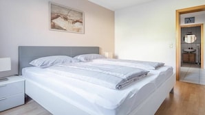 Quarto moderno com cama, mesas de cabeceira, um quadro na parede e um banheiro visível por uma porta aberta.
