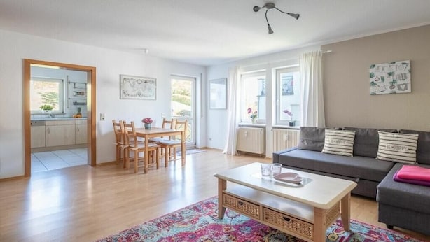 Uma sala de estar com sofá, mesa de centro e mesa de jantar com cadeiras. É visível uma cozinha através de uma porta aberta.