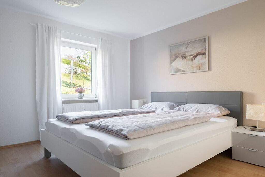 Quarto com cama, mesa de cabeceira, janela com cortinas e um quadro na parede.