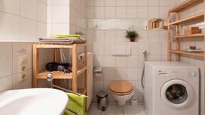 Casa de banho compacta com lavatório branco, prateleiras de madeira, sanita, máquina de lavar roupa e um pequeno contentor de lixo.