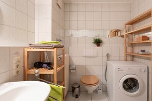 Un bagno funzionale con lavandino bianco, mensole in legno, un water, una lavatrice e un piccolo cestino.