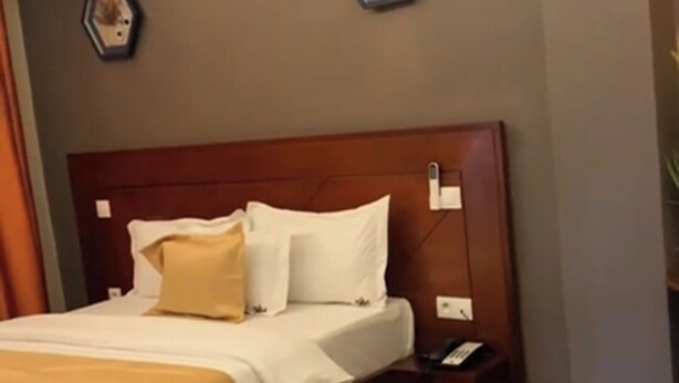 A hotel room with a wooden headboard, a bed with white and beige bedding, a white rug, and a small bedside table with a phone.