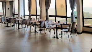 A spacious dining area with multiple tables and chairs, large windows with curtains, and a view of the sea.