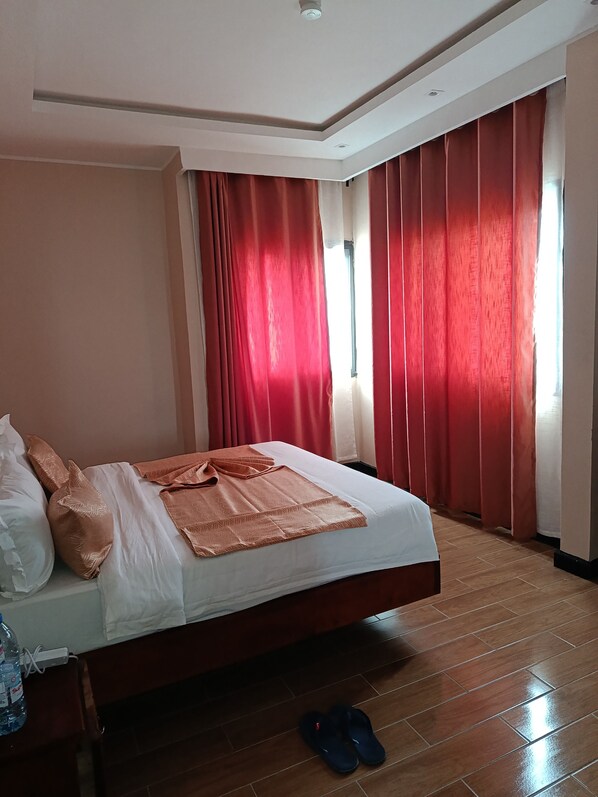 A hotel room with a bed, red curtains, a window, and a pair of slippers on the floor.