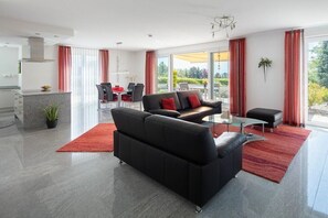 A modern living room with a black sofa, a glass coffee table, and a red rug. There is a dining area with a table and chairs, and a kitchen area with a countertop and cabinets. - Holiday apartment with 1 bedroom (Überlingen)