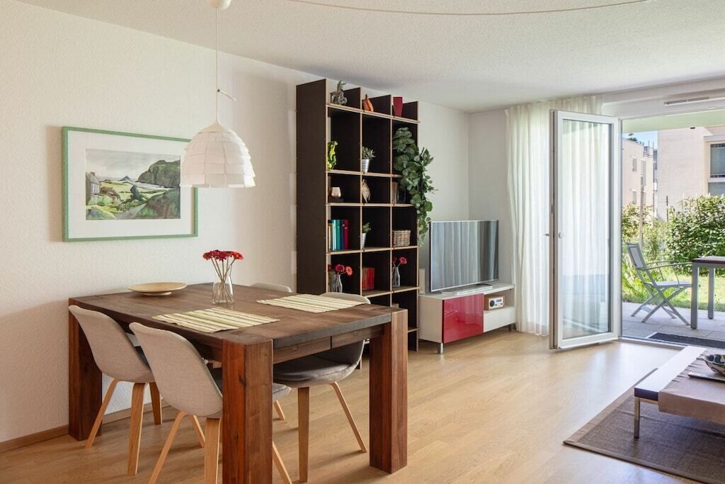 A modern living room with a wooden dining table, chairs, a bookshelf, a television, and a sliding glass door leading to an outdoor area.