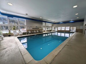 An indoor swimming pool with clear blue water, surrounded by white chairs and potted plants. - Lake View | Top Floor | Shared Pool (Keystone)