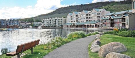 A lakeside walkway with a bench, large rocks, and a row of multi-story buildings with green roofs.
