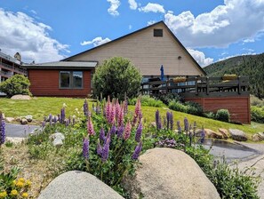 A two-story building with a sloped roof, a wooden deck, and a garden with purple flowers and rocks. - Lake View | Top Floor | Shared Pool (Keystone)