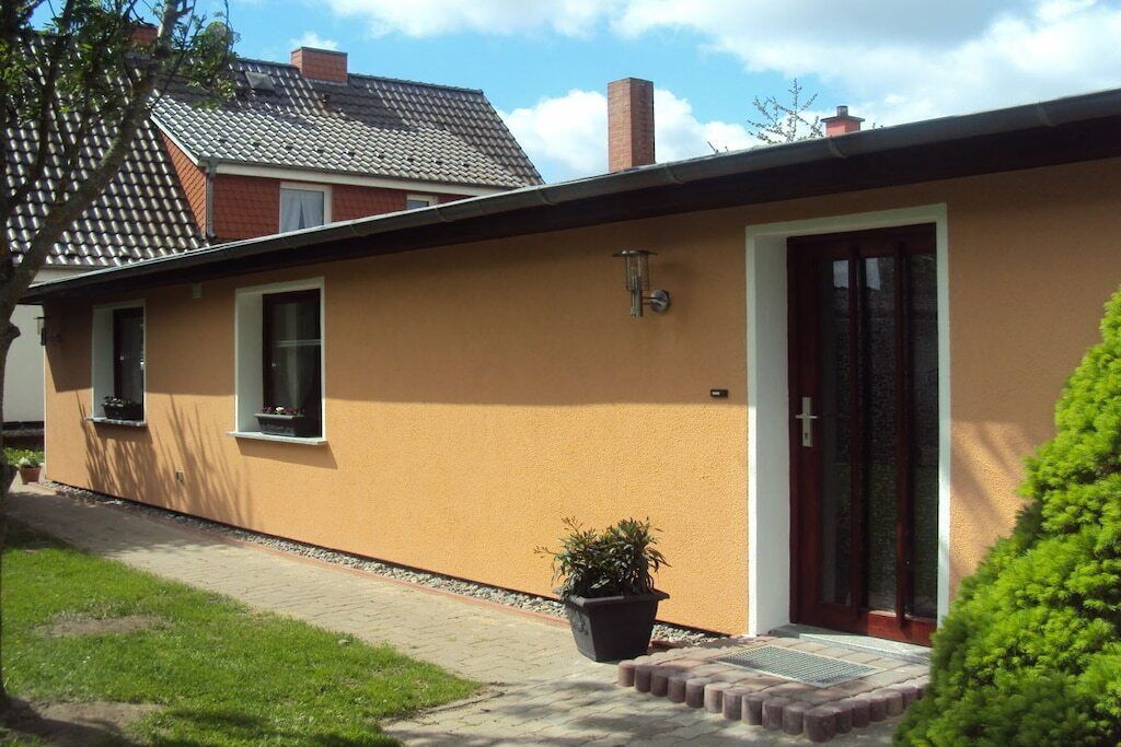 Une maison de plain-pied à l’extérieur brun, avec une porte d’entrée et un petit jardin devant, où se trouve un pot de fleurs.