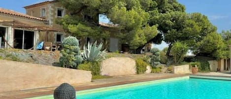 A swimming pool with a wooden deck, surrounded by greenery and a building with a tiled roof.