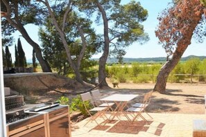 An outdoor patio with a grill, dining table, and chairs, surrounded by trees and a view of the countryside. - Gîte Pomme de Pin 2/4 personnes (LEZIGNAN CORBIERES)