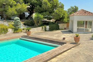 A swimming pool with a wooden deck, surrounded by various plants and trees. - Gîte Pomme de Pin 2/4 personnes (LEZIGNAN CORBIERES)