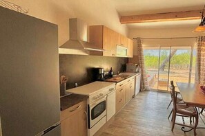 A kitchen with a stove, oven, and a dining area with a table and chairs. - Gîte Pomme de Pin 2/4 personnes (LEZIGNAN CORBIERES)