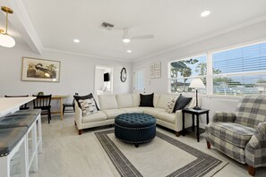 A modern living room with a sectional sofa, ottoman, and a plaid armchair. There is a dining area with a table and chairs, a ceiling fan, and a window with blinds. - Venice Island Florida pool Home (Venice)