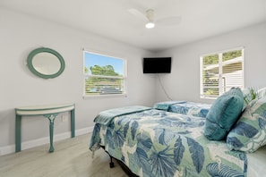 Une chambre à coucher comprenant un lit, une table de chevet, un ventilateur de plafond, un téléviseur fixé au mur et deux fenêtres équipées de stores.