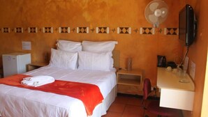 Quarto de hotel com cama, escrivaninha, cadeira, televisão, ventoinha e pequeno frigorífico.
