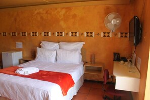 A hotel room with a bed, a desk, a chair, a television, a fan, and a small refrigerator. - Room-thornycroft Lodge & Spa Kugompo (East London)