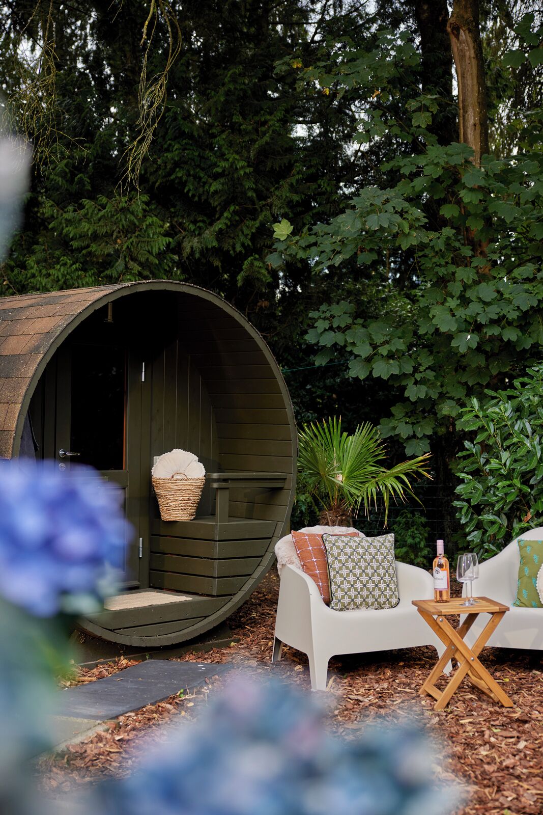 A rustic outdoor seating area with a large wooden barrel structure, a white chair with patterned cushions, and a small table with a bottle and glass.