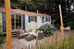 A cozy outdoor seating area with a sofa and chairs, a wooden fence, and a large bush with hydrangeas. - 4P Modern Chalet with Private Sauna - Fenced Forest Garden (Putten)