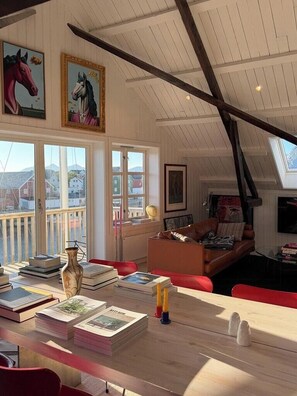 A cozy room with a sloped ceiling, a wooden table with books and a vase, a red sofa, and a window with a view of buildings. - Top-floor flat by the harbour in Henningsvær (Henningsvær)