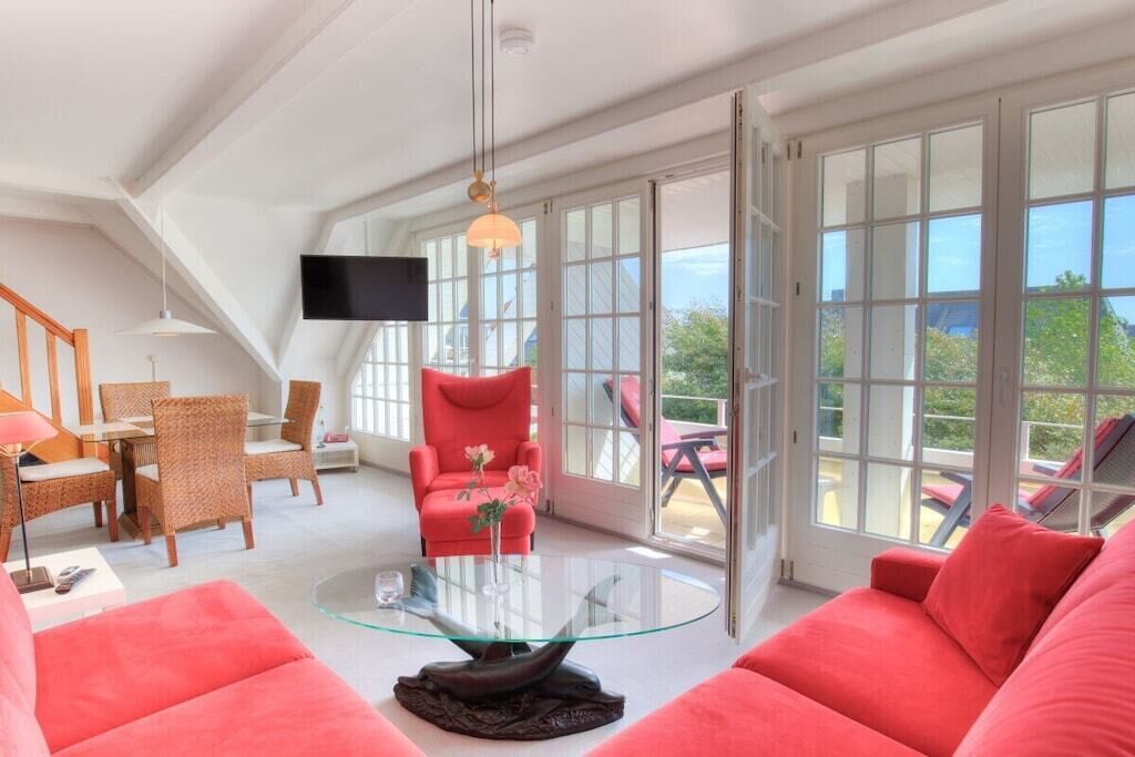 Un vaste espace de vie agrémenté d’une table basse en verre, de canapés rouges, de fauteuils en osier, d’un téléviseur à écran plat et de grandes fenêtres offrant une vue sur l’extérieur.