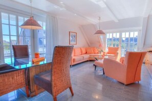 A modern living room with a glass dining table, wicker chairs, a sofa, and orange cushions. - Holiday apartment in Sylt with 1 bedroom (Wenningstedt)