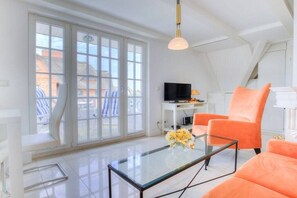 En moderne stue med et glasbord, orange lænestole, et fladskærms-tv og en stor skydedør til et udendørs område.