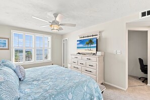 Ein Schlafzimmer mit einem Bett, einem Deckenventilator, einem Fernseher auf einer Kommode und einem Fenster mit Jalousien.