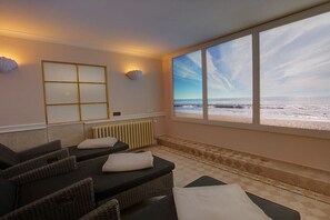 Une salle de spa dotée d’une grande fenêtre donnant sur une plage, d’une table de massage et d’un luminaire fixé au mur.