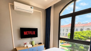 Hotellihuone, jossa on sänky, televisio, jossa näkyy Netflix, ikkuna, josta on näkymä rakennuksiin, ja seinälle asennettu ilmastointilaite.