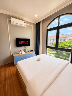 A hotel room with a bed, a television displaying 'NETFLIX,' a window with a view of buildings, and a wall-mounted air conditioner. - InaVay Retreat | 2 mins walk to Beach | Heart of Grand World, Phu Quoc (Phu Quoc)