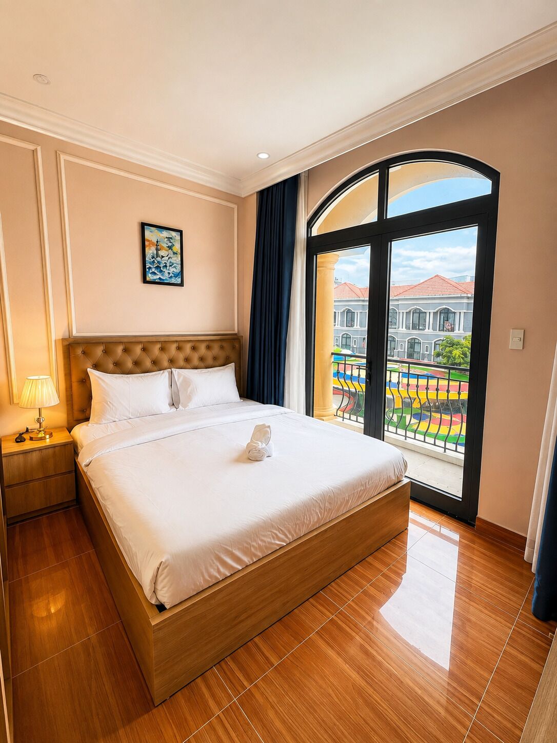 A hotel room with a large bed, a nightstand, a painting on the wall, and a view of a colorful outdoor area through a large window.