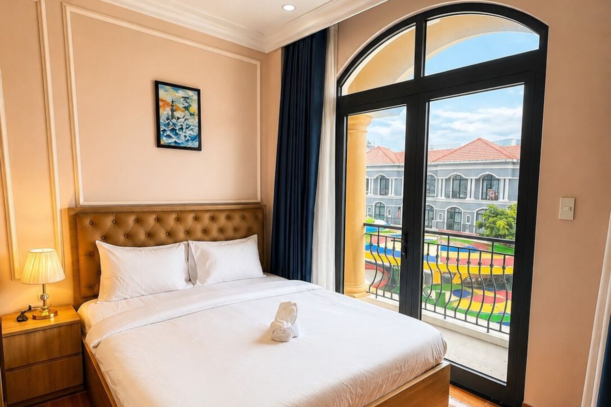 A hotel room with a large bed, a nightstand, a painting on the wall, and a view of a colorful outdoor area through a large window.
