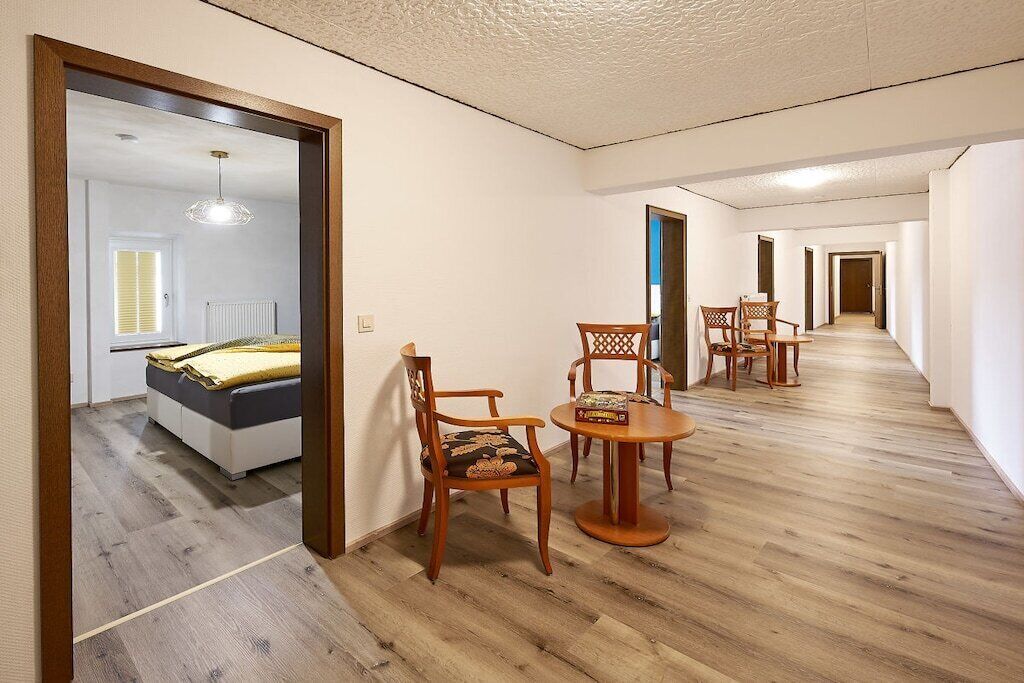 Une chambre d’hôtel avec un plancher en bois, un lit recouvert d’une couverture jaune, une petite table avec des chaises et un couloir menant à d’autres chambres.
