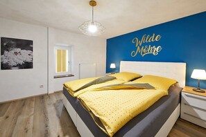 Een slaapkamer met een bed, een nachtkastje, een lamp, een muur met een bord, een raam met zonwering en een radiator.