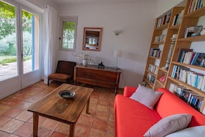 Ein Zimmer mit einem roten Sofa, einem Holztisch, einem Sessel, einem Bücherregal und einem Fenster mit Vorhängen.