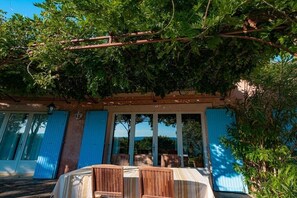 Een overdekt terras met houten meubels, een pergola erboven en uitzicht op een gebouw met blauwe luiken.