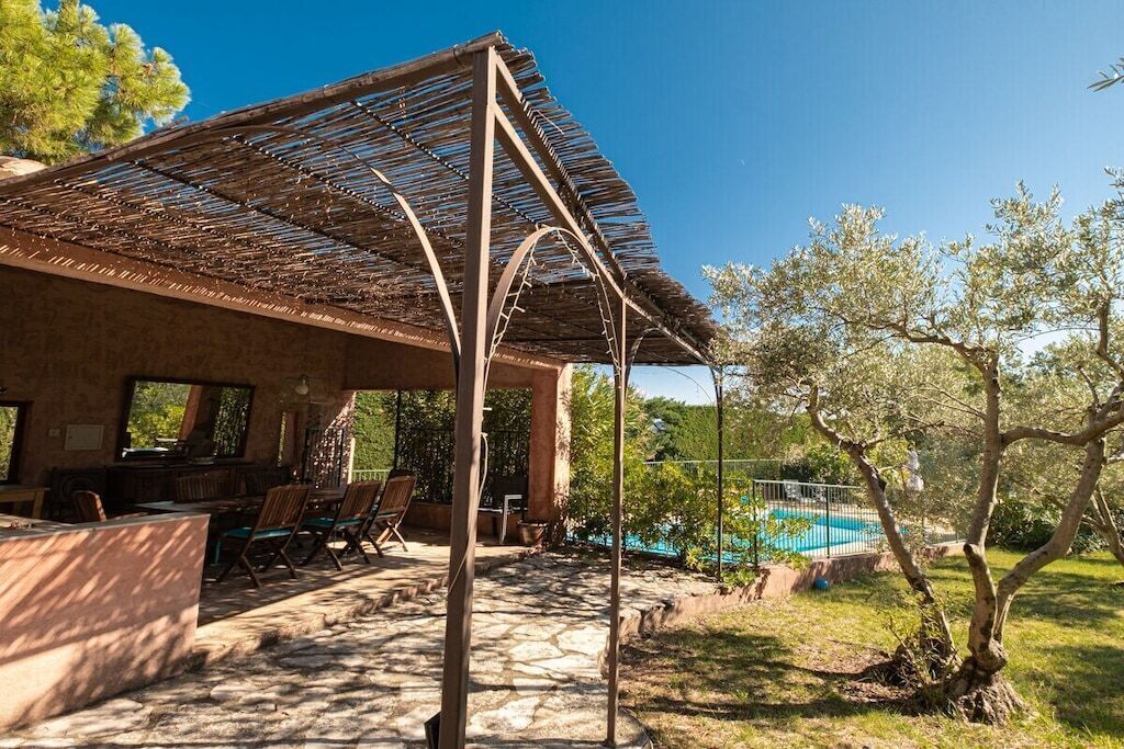 Un'area relax all'aperto coperta, con pavimento in pietra, una piscina sullo sfondo e alberi tutt'intorno.