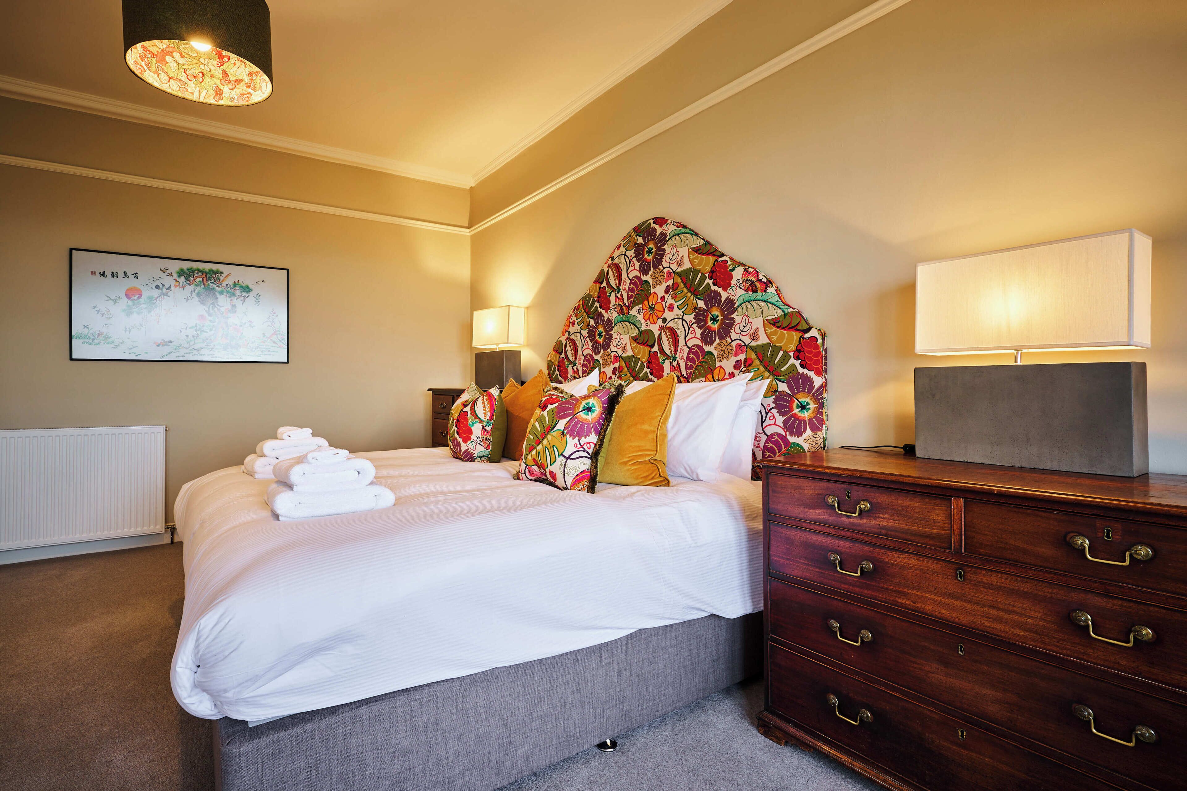 A hotel room with a neatly made bed, a dresser with drawers, a bedside lamp, and a wall-mounted lamp.