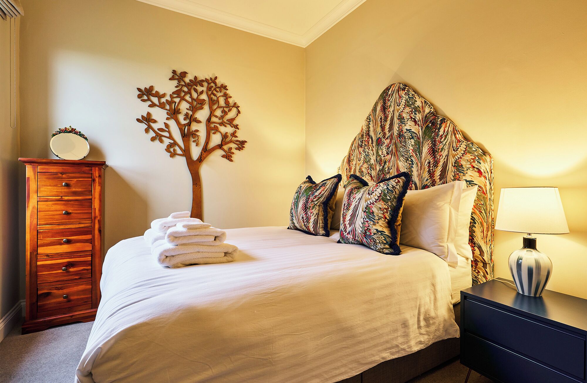 Una cama bien hecha con un cabecero con forma de árbol, un aparador de madera y una lámpara en una mesita de noche.