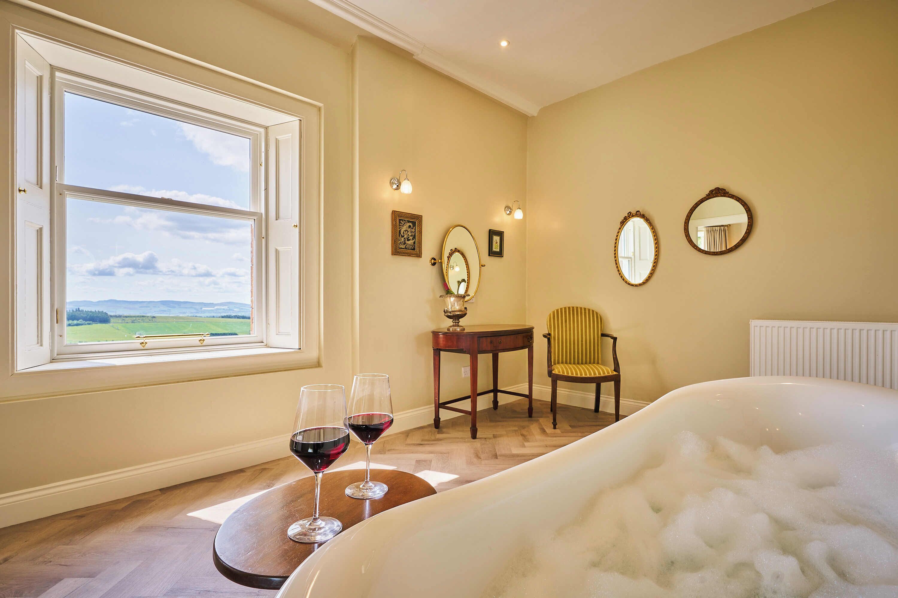 A room with a large window, a bed, a chair, and two glasses of red wine on a small table.