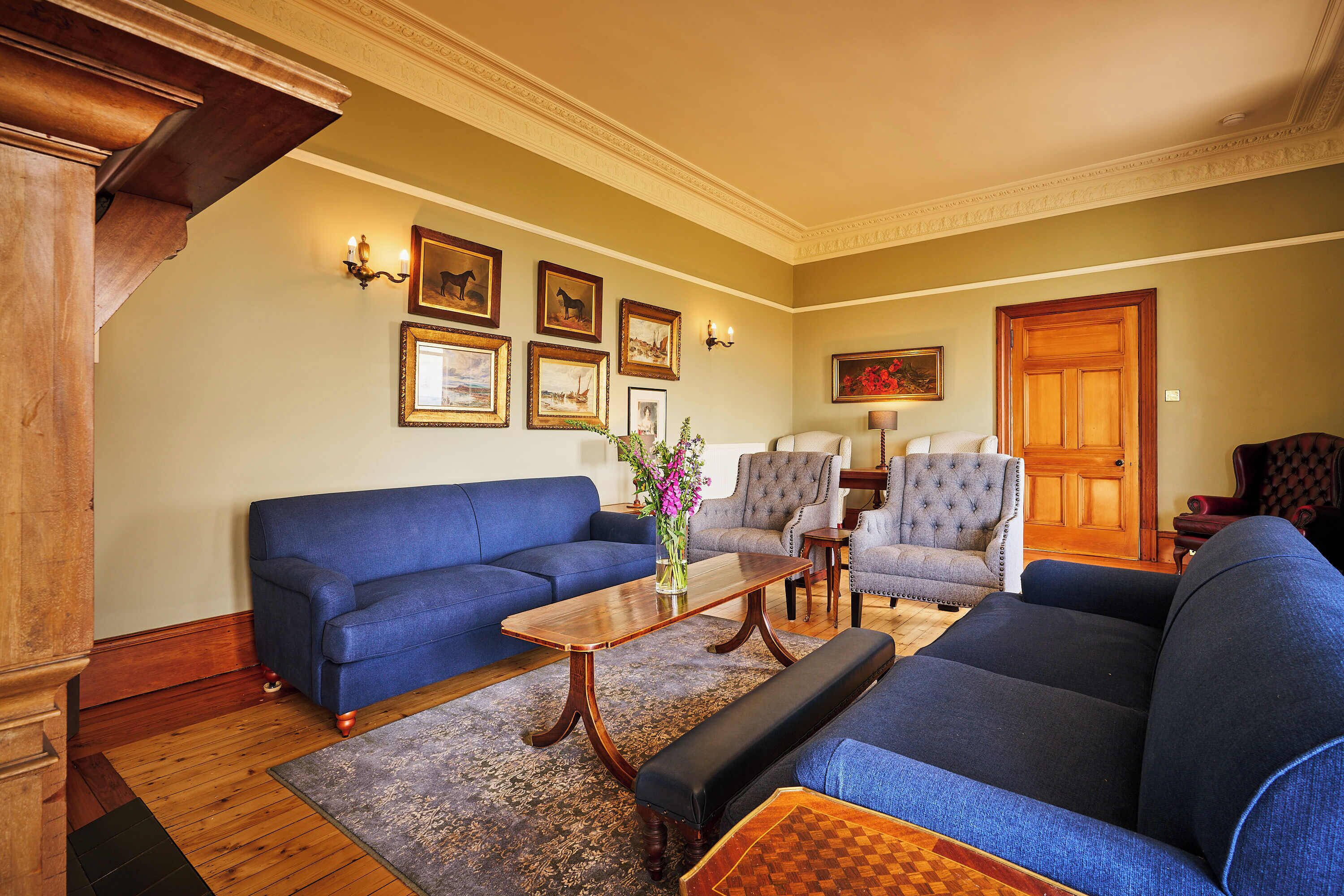 Una sala de estar con un sofá azul, una mesa de centro de madera y un jarrón floral. Hay cuadros en la pared y una puerta de madera.