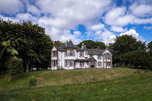 A large, two-story house with a prominent turret and multiple windows, surrounded by a grassy area and trees.