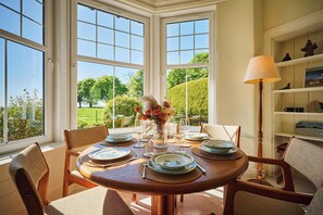 Comedor con una mesa redonda dispuesta para cuatro personas, sillas alrededor y vista a un jardín a través de amplias ventanas.