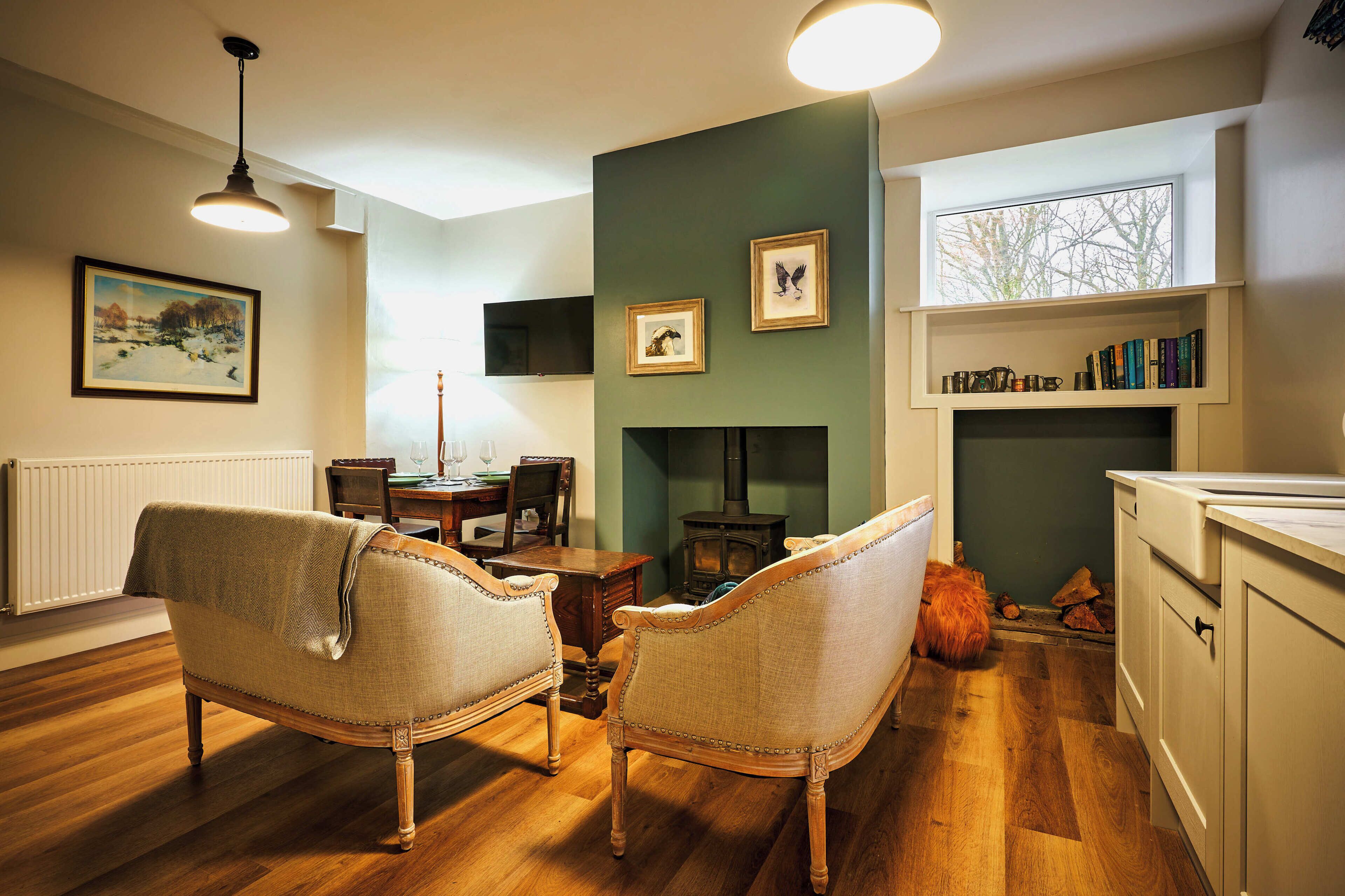 A cozy living room with wooden flooring, a fireplace, a dining table with chairs, and a bookshelf.